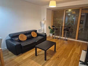 A modern living room with a black leather sofa, a wooden coffee table, a floor lamp, and a large window with blinds.