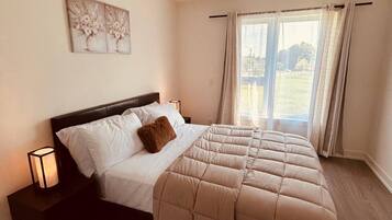 A bedroom with a large bed, a nightstand, a wall-mounted lamp, and a window with curtains.