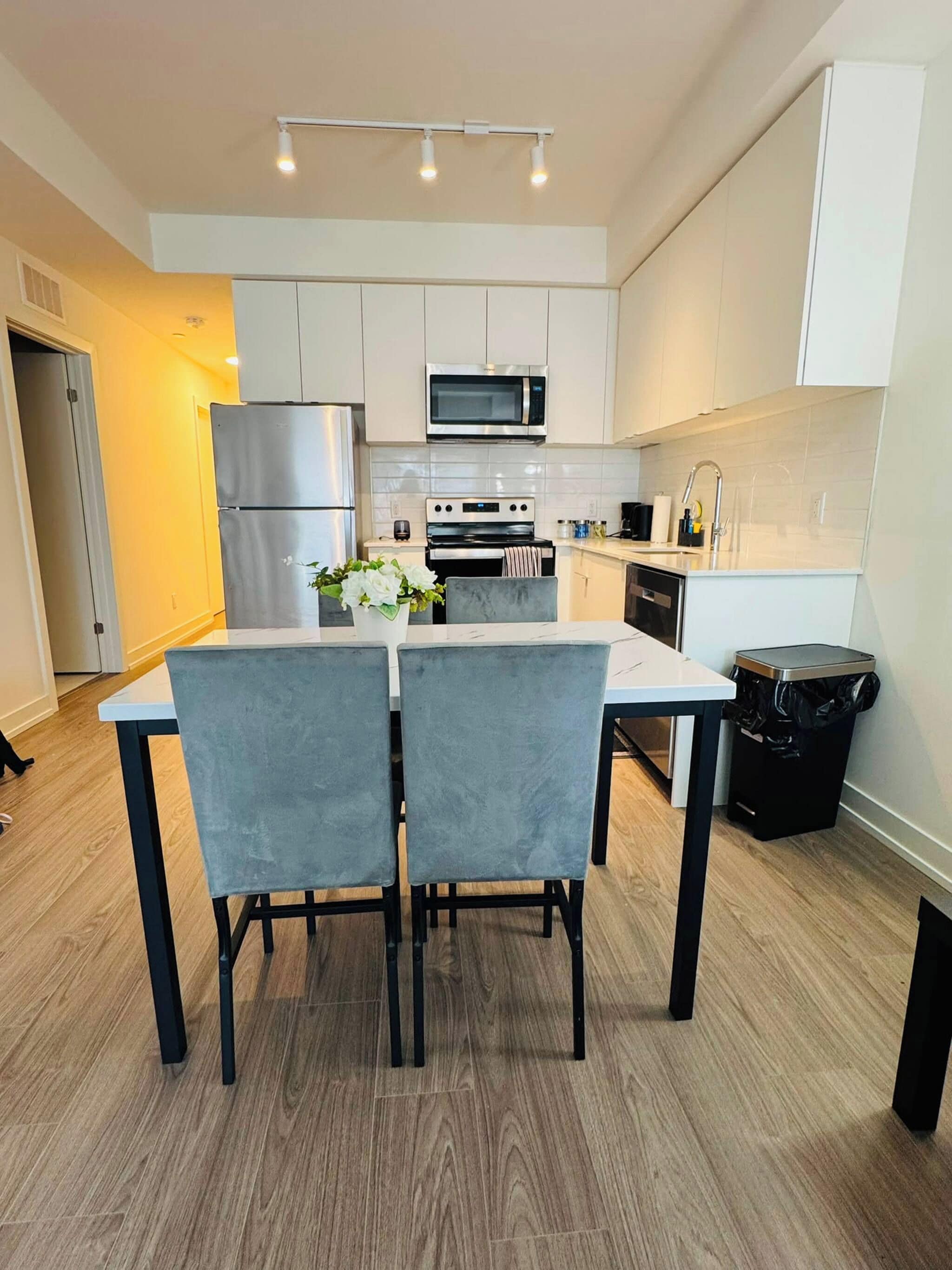 A modern kitchen with white cabinets, stainless steel appliances, a dining table with chairs, and a trash bin.