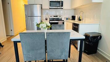 A modern kitchen with white cabinets, stainless steel appliances, a dining table with chairs, and a trash bin.
