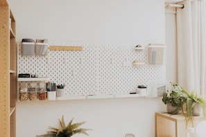 Eine weiße Wand mit einem Gitterhaken-System, Ablageflächen und Regalen, ein kleines Holzregal mit Büromaterialien und ein Blumentopf auf einem Beistelltisch.