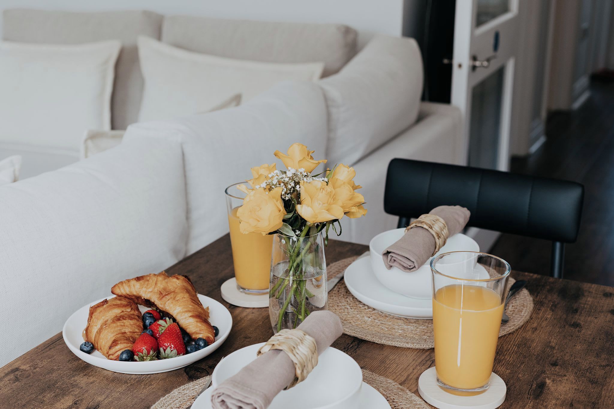 A breakfast setting with croissants, strawberries, blueberries, a glass of orange juice, and a vase with yellow flowers on a wooden table.