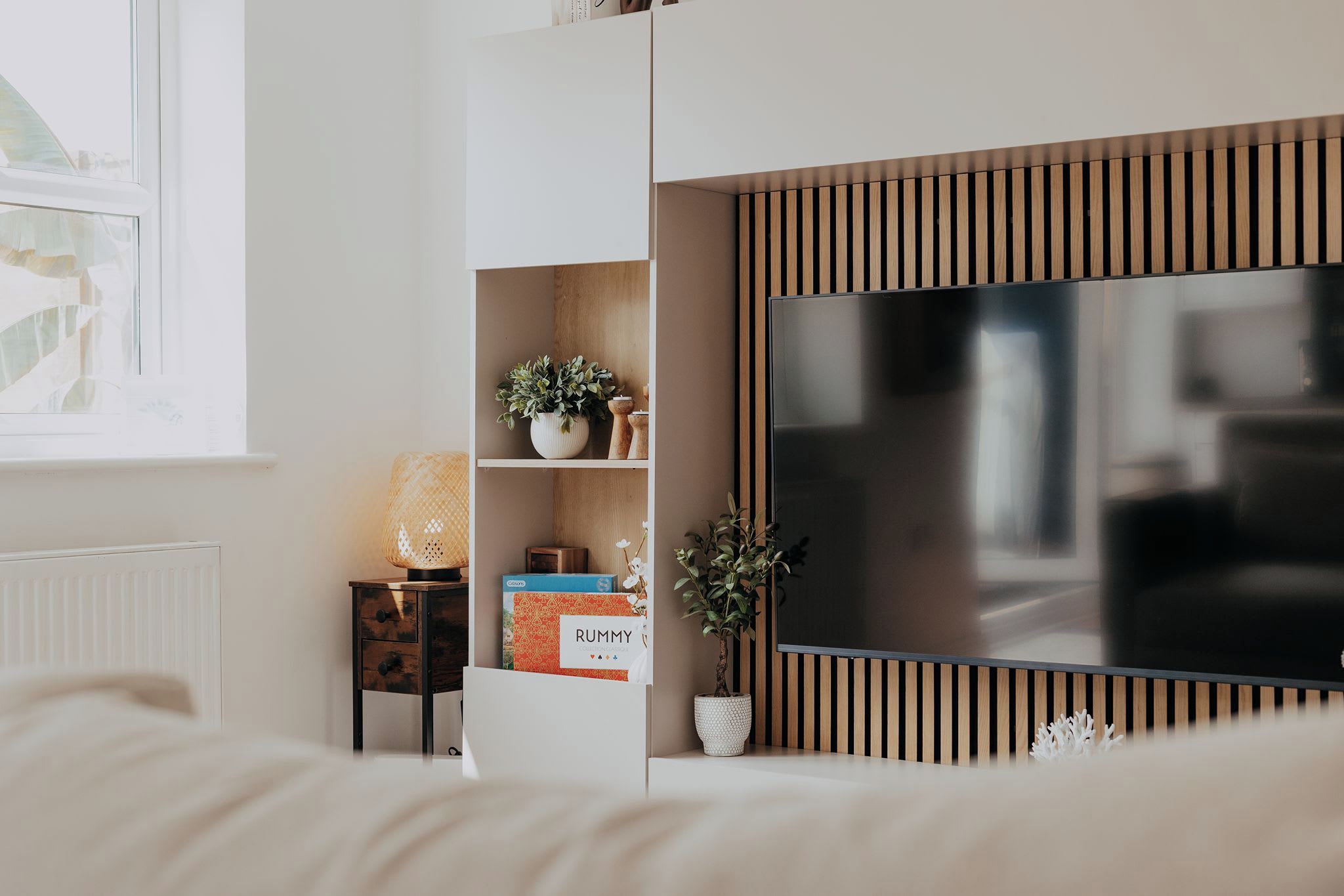 A modern bedroom with a bed, a TV, a shelf with decorative items, and a window.
