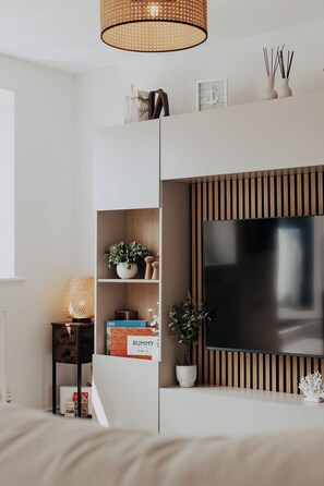 A modern living room with a flat-screen TV mounted on a wooden panel wall, a white shelving unit with decorative items, and a wicker lamp. - Coastal Hideaway by Lil Blu Stays (Bournemouth, Christchurch and Poole)