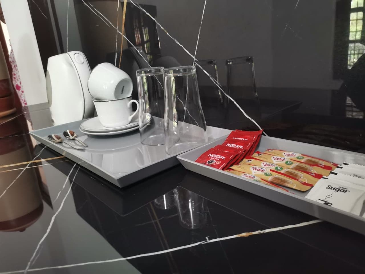 Une chambre d’hôtel moderne avec une table en marbre noir, une machine à café blanche, des verres transparents, un plateau avec des dosettes de café et un sachet de sucre.