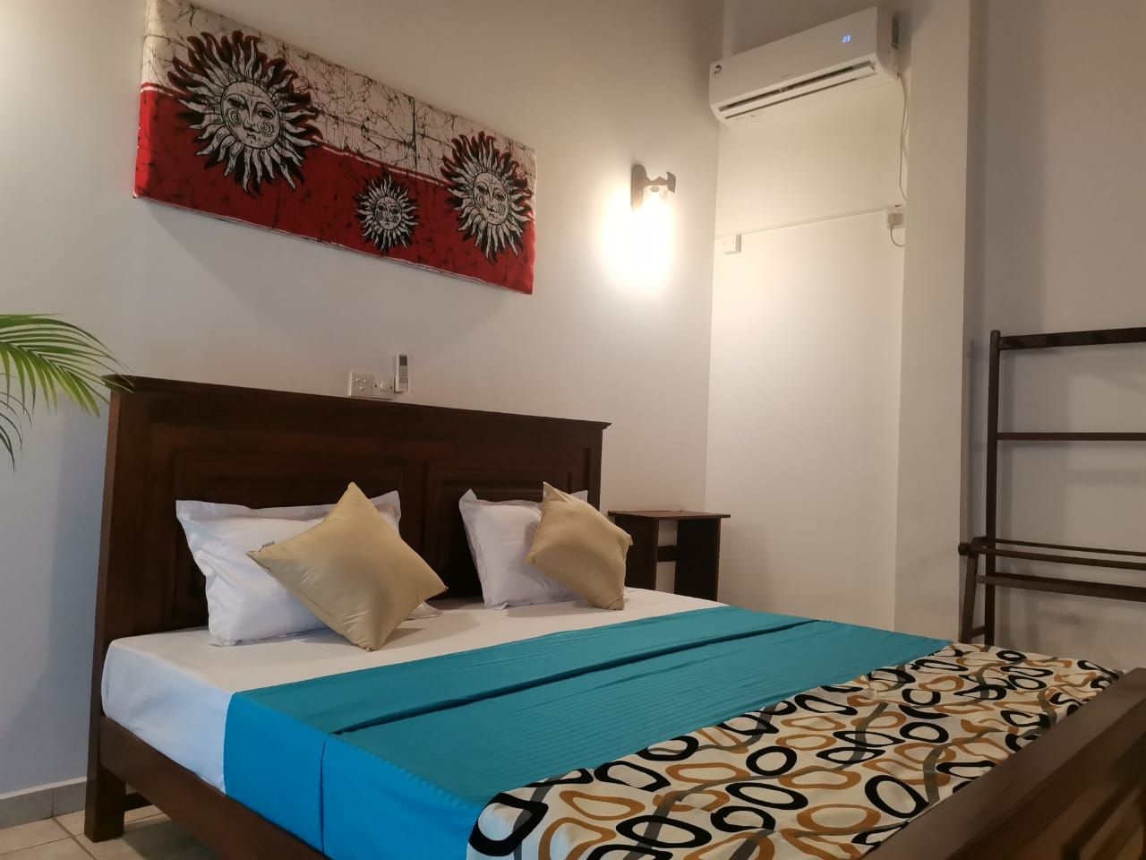 A hotel room with a bed, a wall-mounted air conditioner, a wall art piece, and a bedside table.