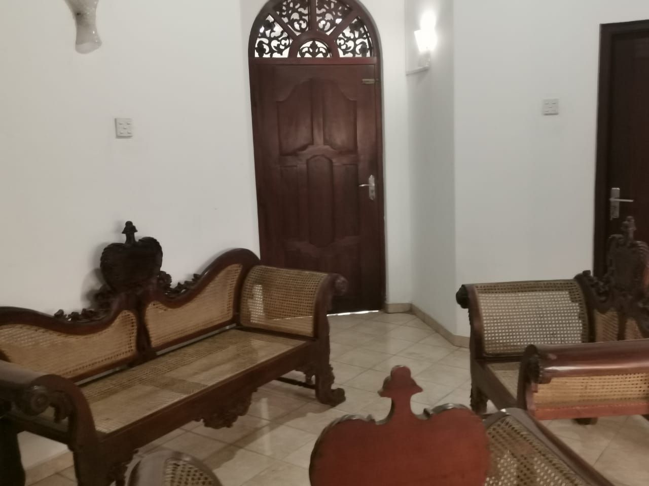 A room with two wooden sofas with woven seats, a dark wooden door with intricate carvings, and a tiled floor.