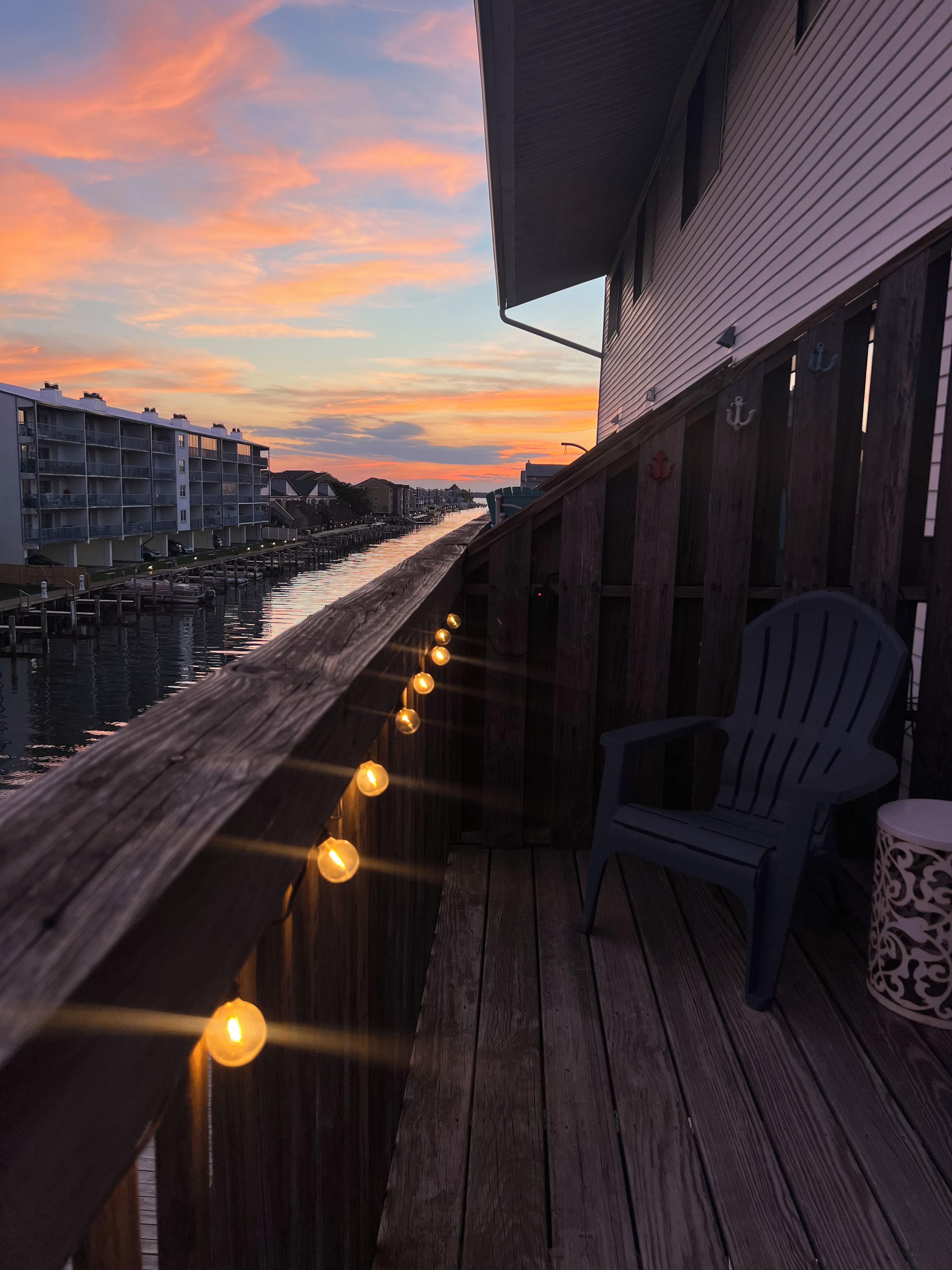A wooden deck with a row of lights, a chair, and a view of a waterway with buildings in the background.