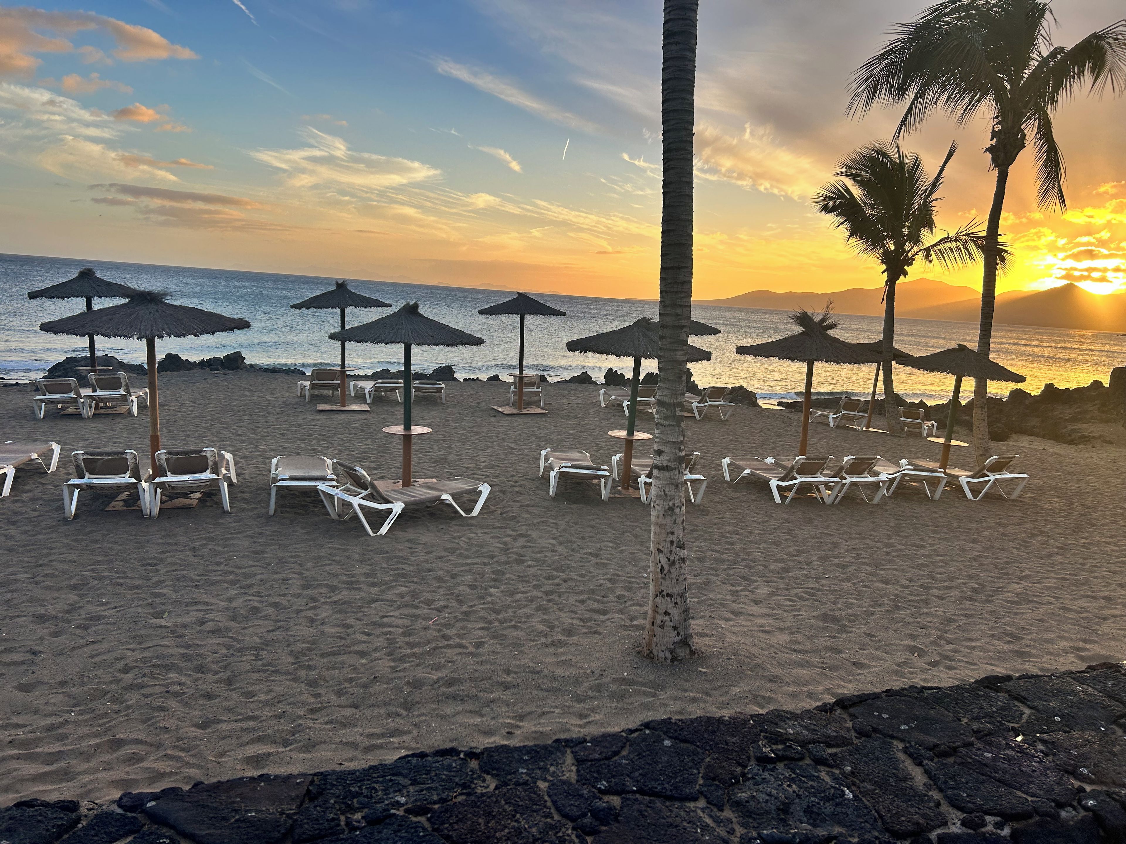 Une plage avec des transats et des parasols en chaume, un ciel dégagé et un coucher de soleil sur l’océan.