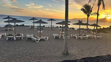 Une plage avec des transats et des parasols en chaume, un ciel dégagé et un coucher de soleil sur l’océan.