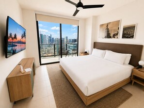 Ein Hotelzimmer mit einem großen Bett, einem Schreibtisch, einem Sessel, einem Nachttisch, einem Deckenventilator und Blick auf die Stadt durch das Fenster.
