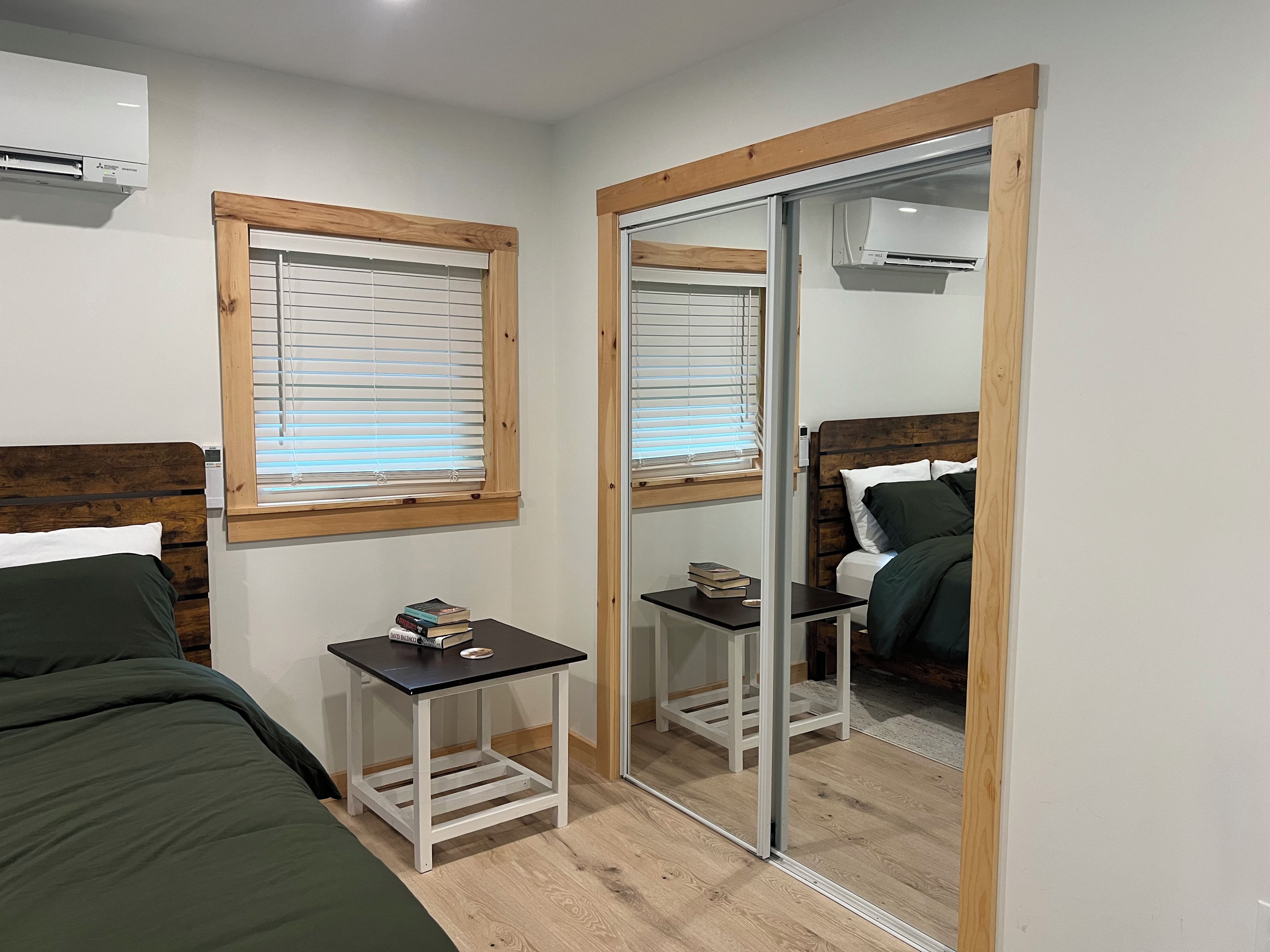 A bedroom with a bed, a bedside table with books, a mirror, and an air conditioning unit.