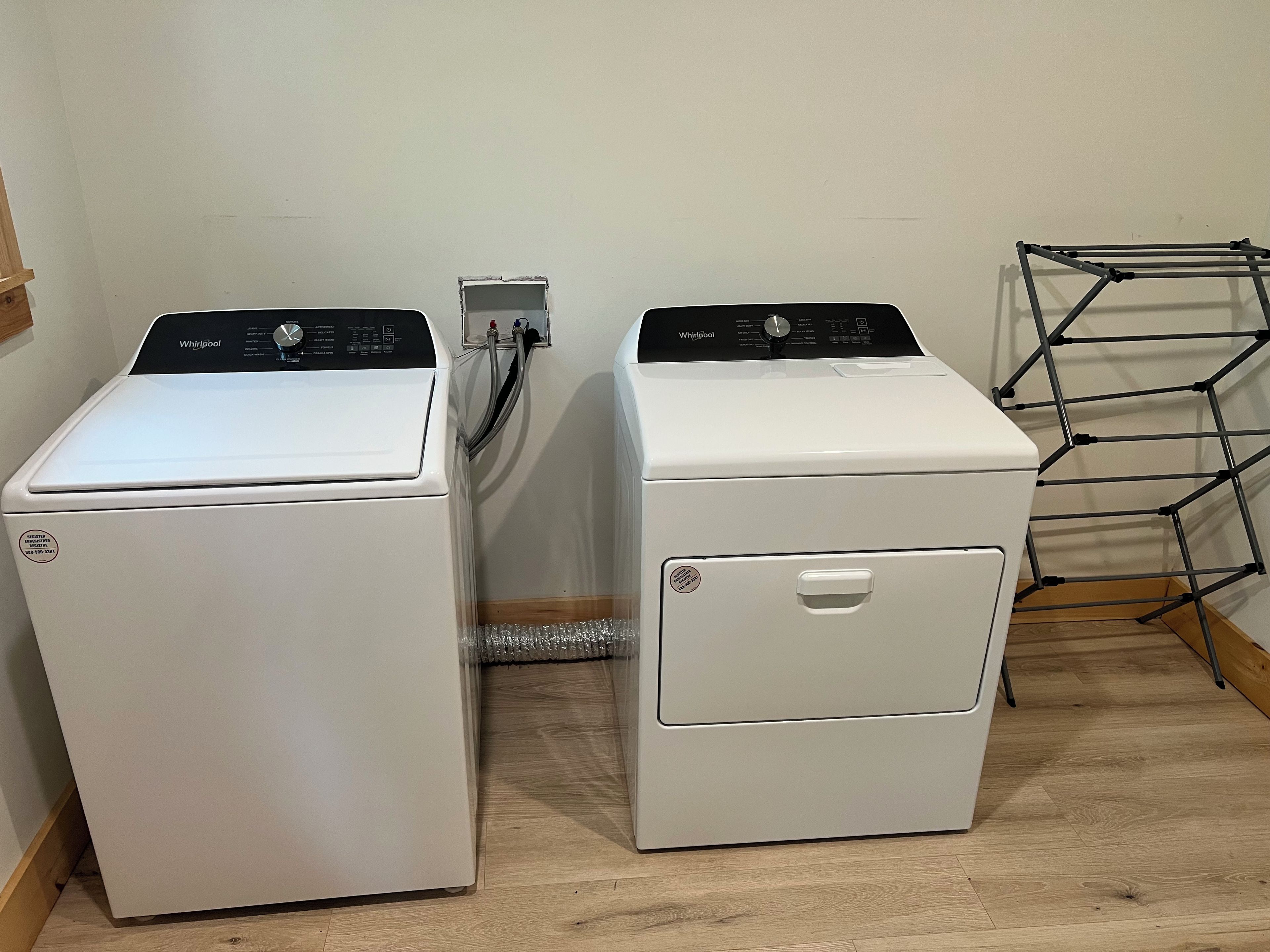 Two white Whirlpool front-loading washing machines in a room with wooden flooring and a metal drying rack.