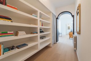 Ein Flur mit weißen Regaleinheiten, gefüllt mit Büchern, einem schwarzen Schreibtisch mit Stuhl und einem großen Spiegel an der Wand.