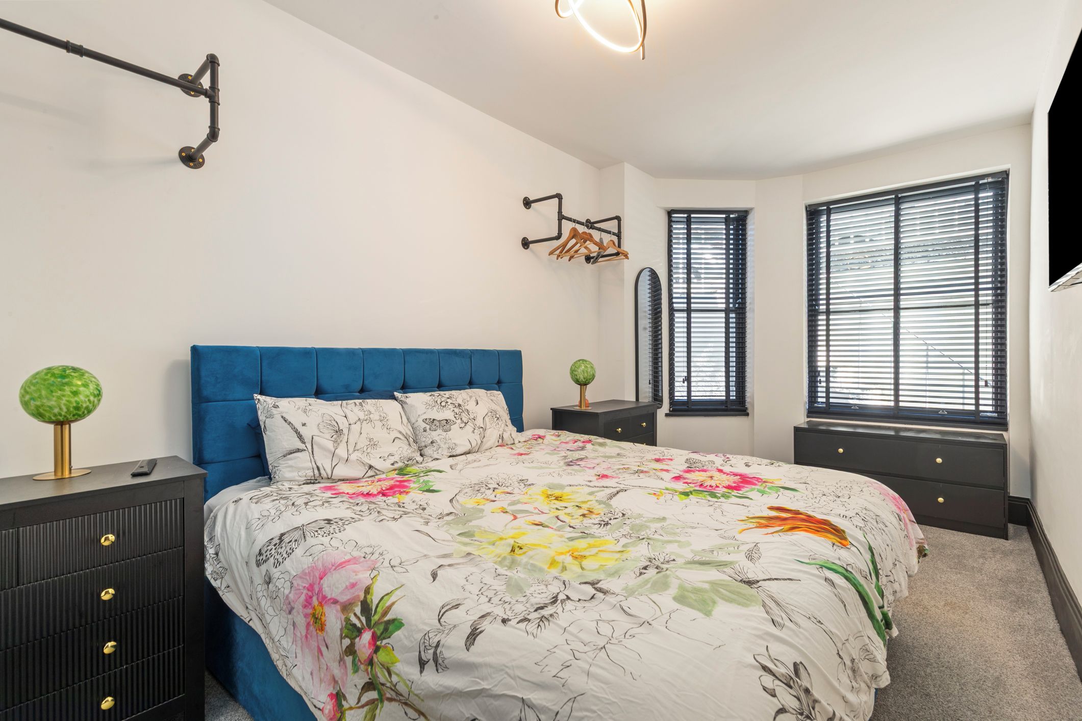 A bedroom with a blue headboard, floral bedding, a dresser with drawers, a mirror, and two windows with blinds.