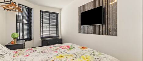 A bedroom with a bed, two windows with blinds, a mounted TV, and a wall-mounted shelf.
