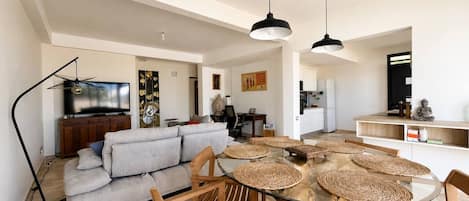 A modern living room with a glass dining table, wooden chairs, a grey sofa, and a TV.
