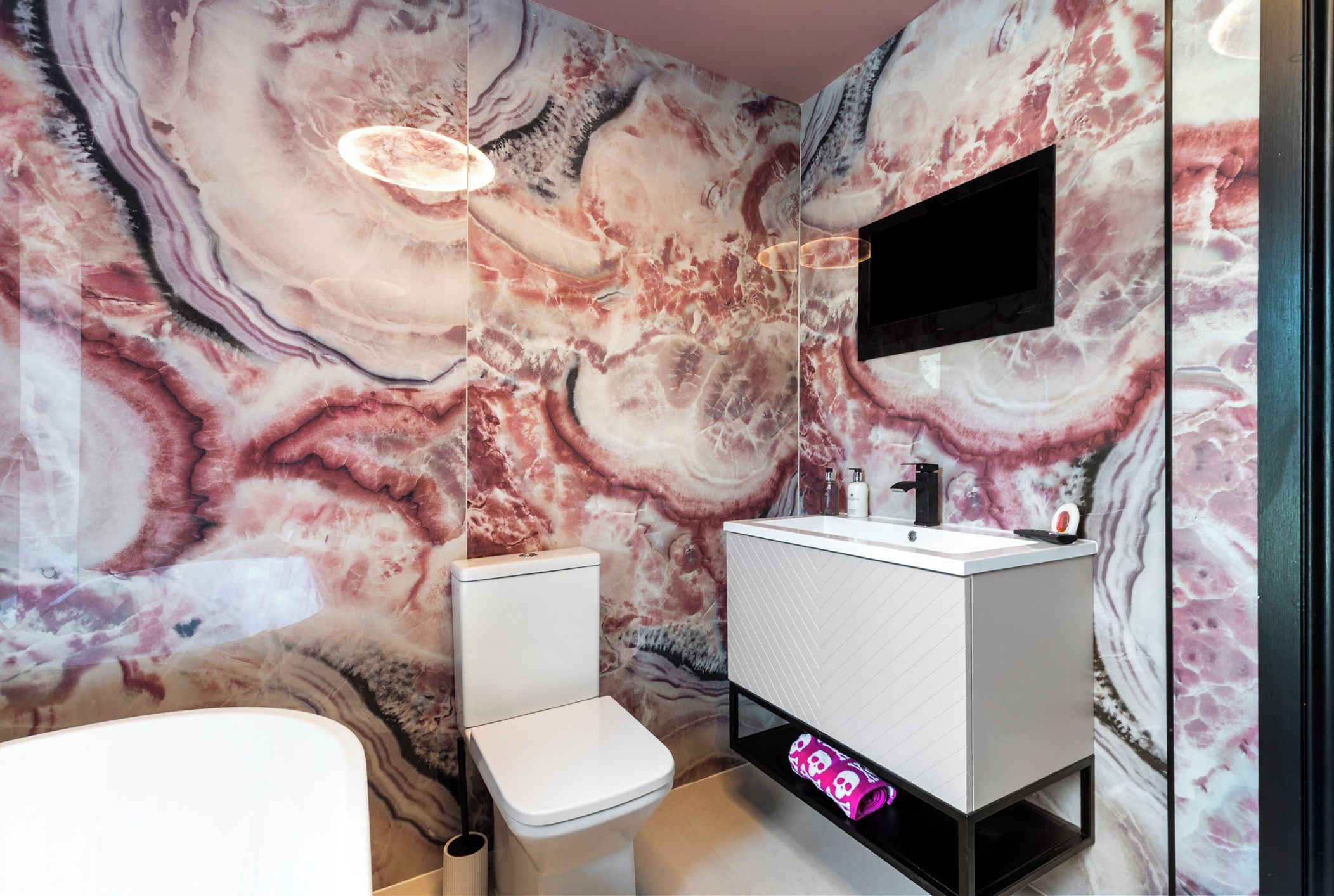 A bathroom with a marble wall, a toilet, a sink, and a TV mounted above the sink.