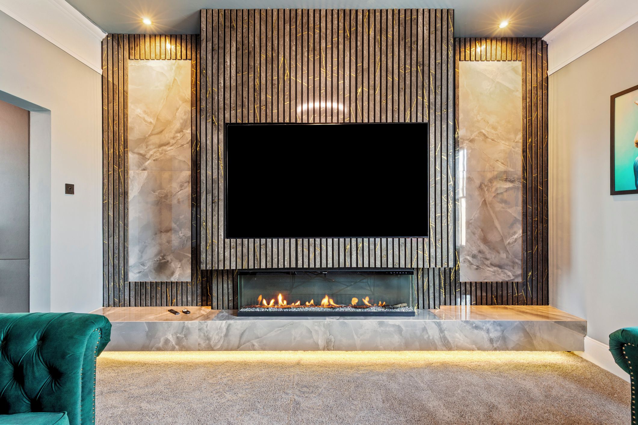 A modern living room with a large flat-screen TV mounted above a fireplace, flanked by marble-like panels, and a carpeted floor.