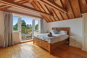 Ein Schlafzimmer mit Holzbalkendecke, einem Bett mit Kissen, einem Nachttisch, einer Kommode und Blick auf einen Balkon mit Gartenmöbeln.