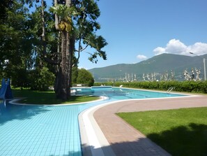 Ein Schwimmbecken mit gefliestem Sonnendeck, umgeben von viel Grün und Blick auf Berge.