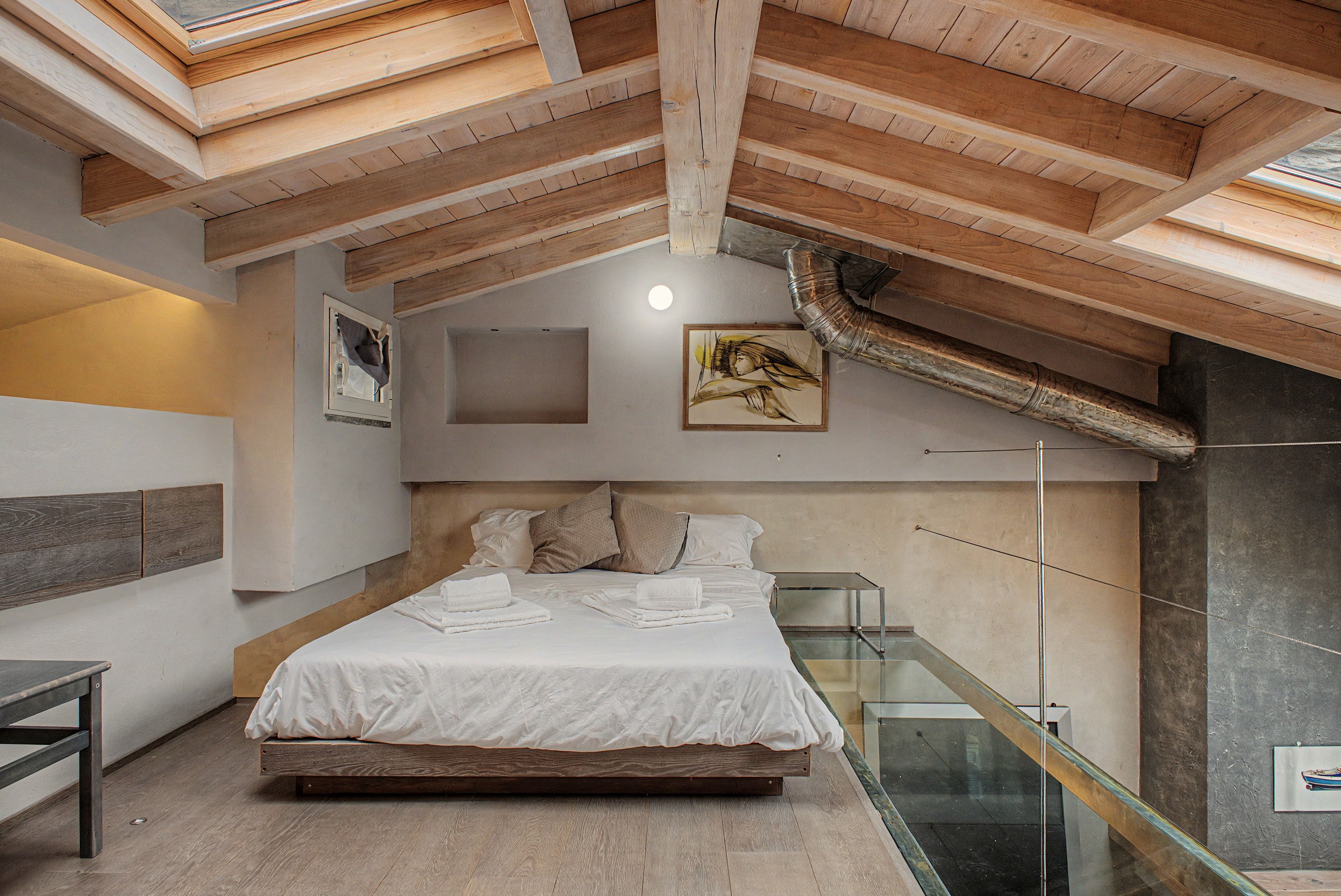 Une chambre avec un plafond en bois, un lit recouvert de draps blancs, une balustrade en verre et un tableau accroché au mur.