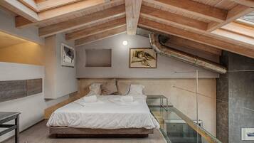 Une chambre avec un plafond en bois, un lit recouvert de draps blancs, une balustrade en verre et un tableau accroché au mur.