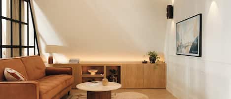 A modern living room with a sofa, a round coffee table, and a small chair. There is a bookshelf with decorative items and a painting on the wall.