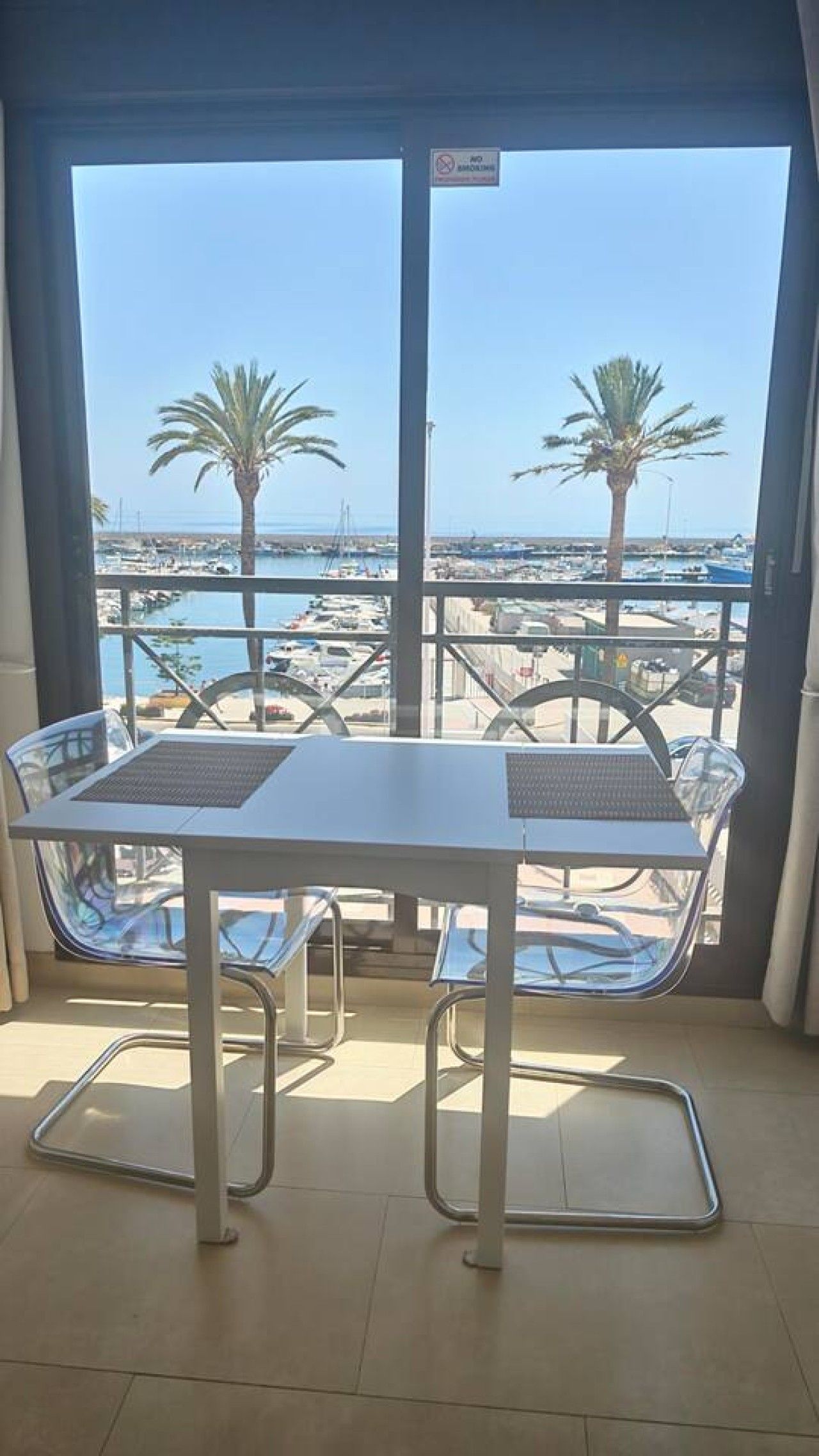 A balcony with a table and chairs, overlooking a marina with boats and palm trees.