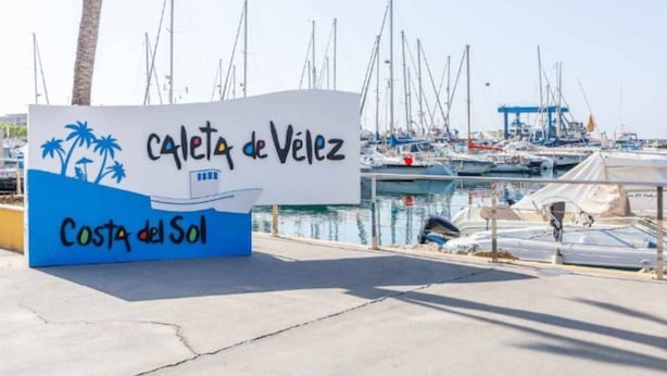 Schild für die 'Calle de Velez' mit einem Yachthafen im Hintergrund.