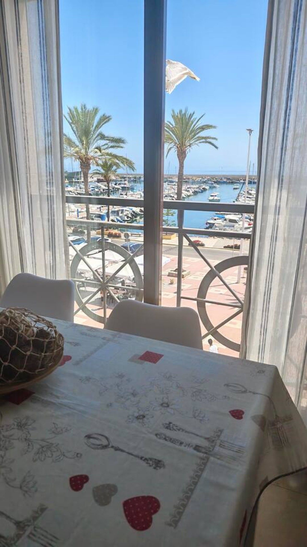 Une table recouverte d’une nappe à motifs, un objet décoratif posé sur la table, et une vue sur une marina avec des bateaux et des palmiers, visible à travers de grandes fenêtres.