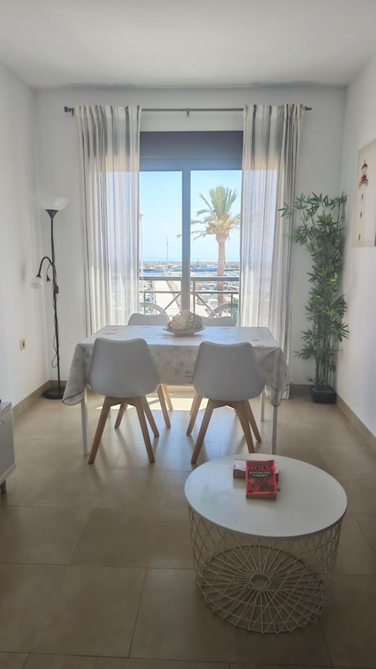A room with a dining table and chairs, a round coffee table with a book, and a view of the sea through large windows.