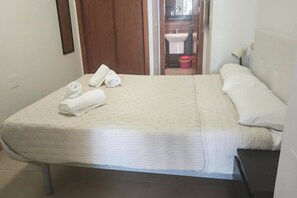 Ein Einzelbett mit weißer Matratze und Kissen, eine Holztür, ein kleines Badezimmer mit Waschbecken und einem Mopp sowie ein gefliester Boden.