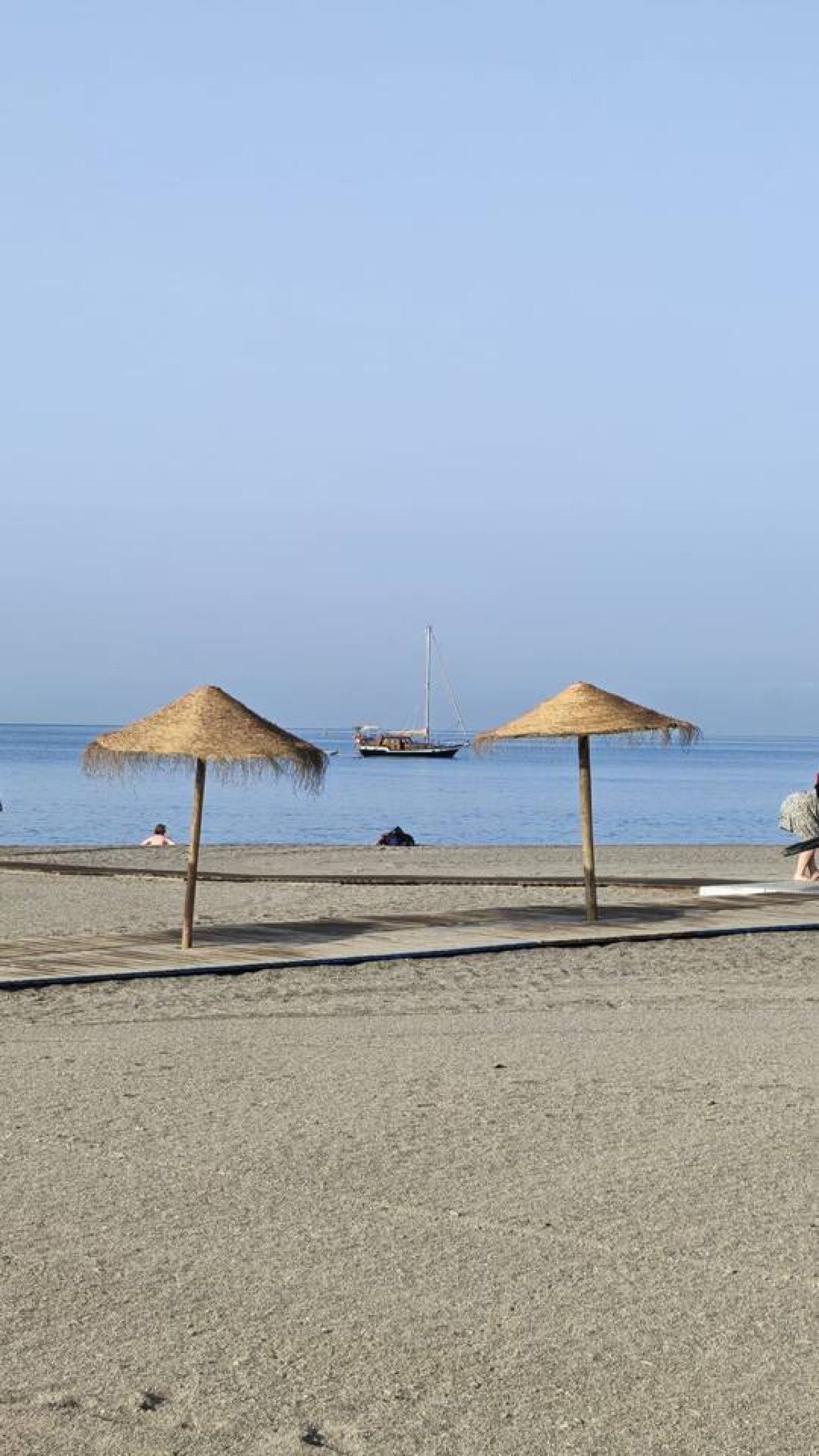 Una playa con dos sombrillas de paja, una orilla arenosa y un velero a lo lejos.