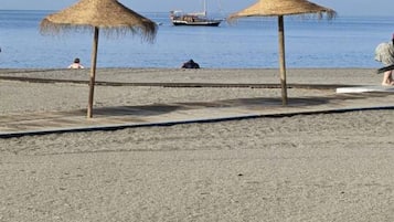 Una playa con dos sombrillas de paja, una orilla arenosa y un velero a lo lejos.