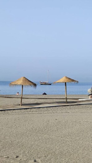 Une plage avec deux parasols en chaume, une rive sablonneuse et un voilier à l’horizon.