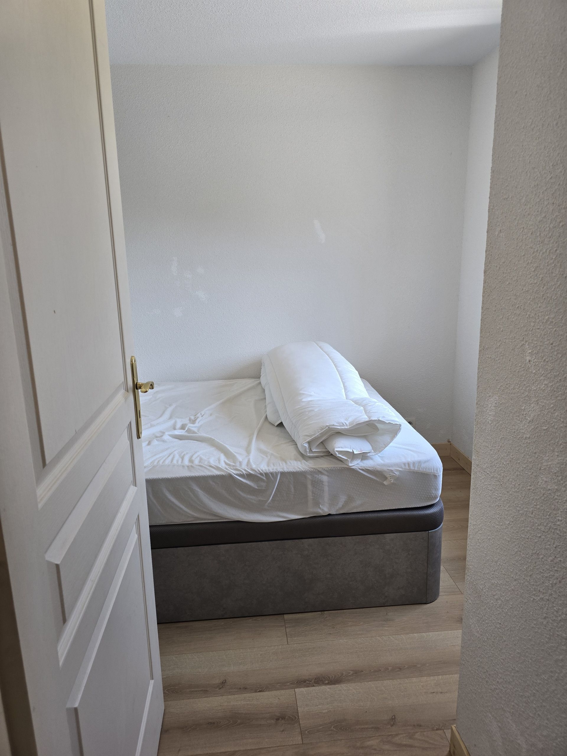 A small, neatly made bed in a room with a wooden floor and white walls.