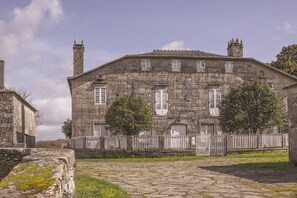 A stone building with a central entrance, flanked by two trees, and a white picket fence. - Pazo Recimil (Lugo)