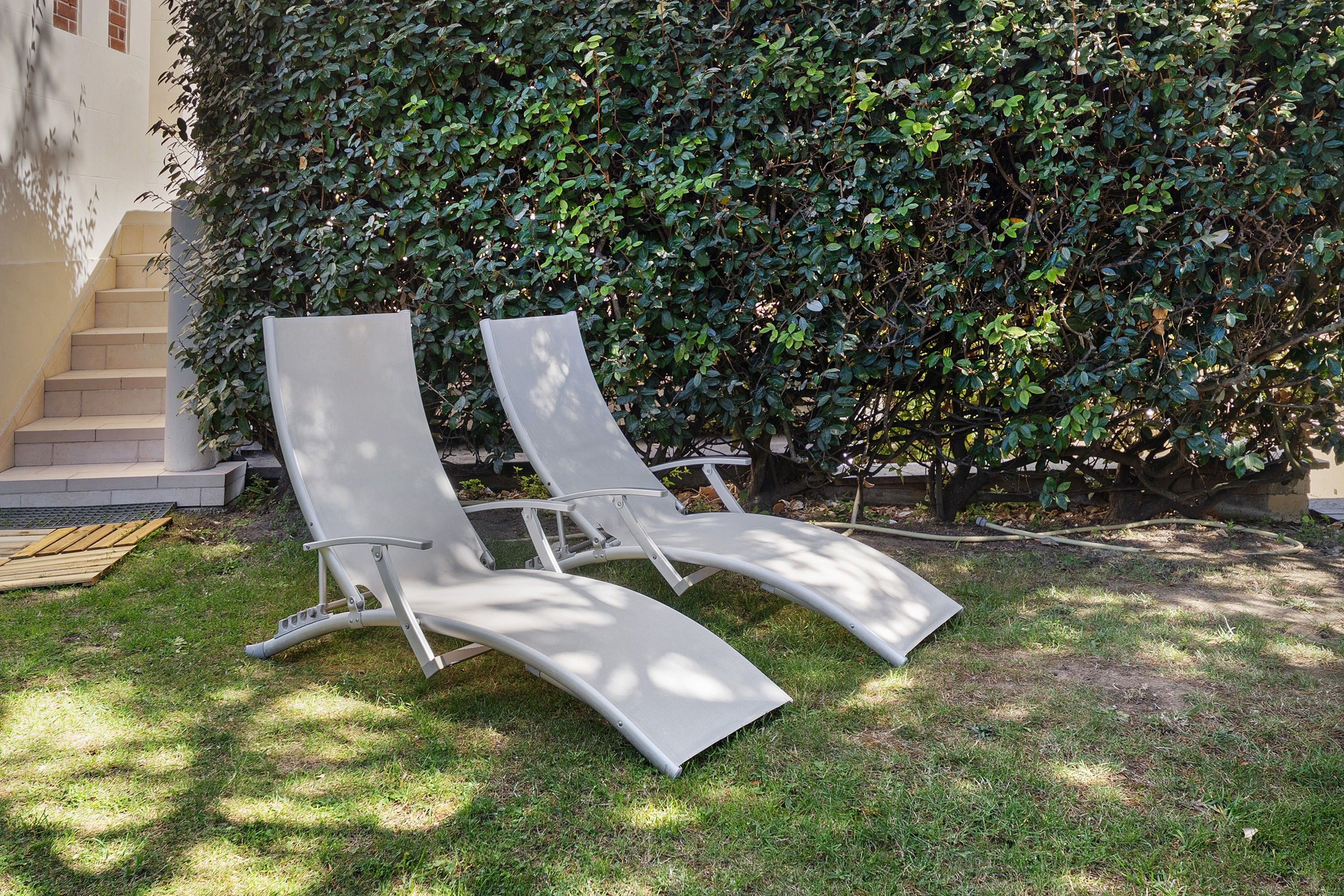 Twee comfortabele stoelen staan op een gazon, met een haag en een gebouw op de achtergrond.