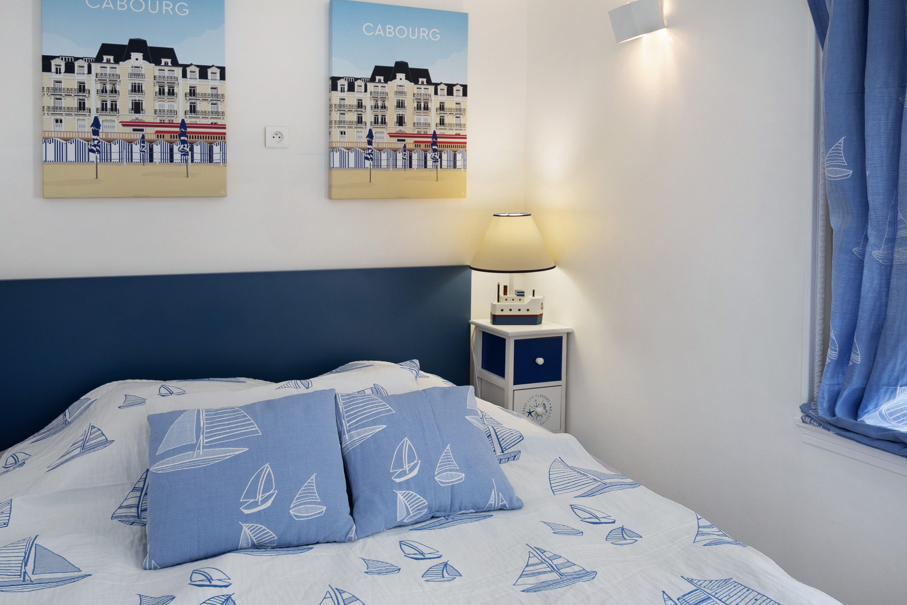Une chambre d’hôtel avec un lit, deux oreillers ornés de motifs de voiliers, une table de chevet avec une lampe et deux affiches représentant une ville côtière.