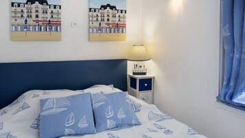 Une chambre d’hôtel avec un lit, deux oreillers ornés de motifs de voiliers, une table de chevet avec une lampe et deux affiches représentant une ville côtière.
