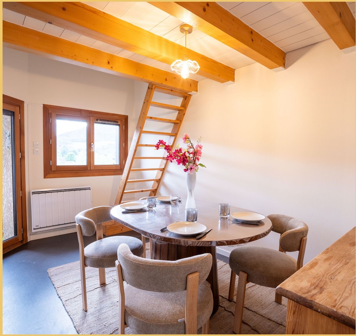 A dining area with a round table, chairs, a vase with flowers, a ladder, and a window.