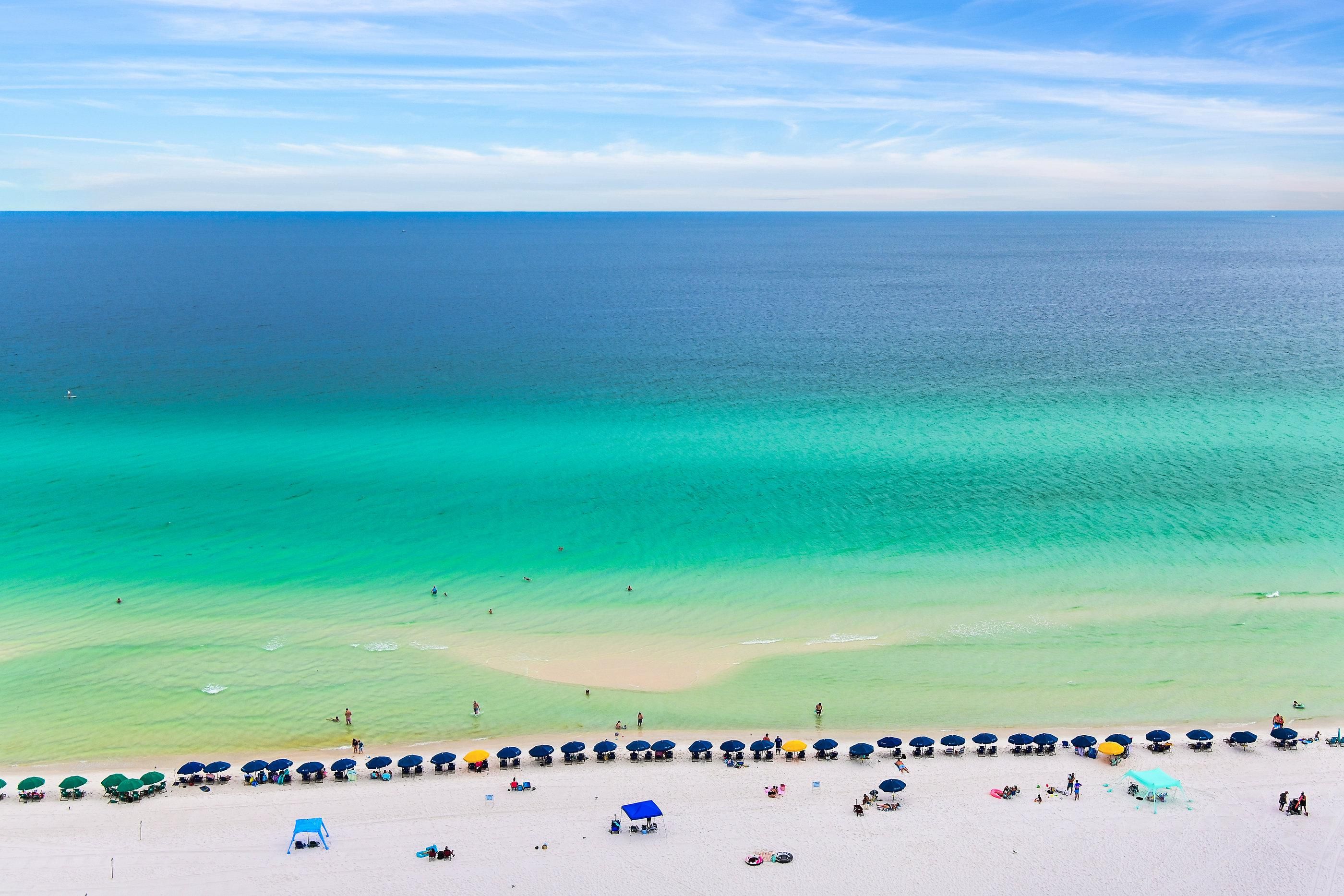 Une plage aux eaux turquoise et limpides, un rivage sablonneux, ainsi que de nombreux parasols et chaises de plage.