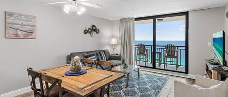 Un coin repas avec une table et des chaises en bois, un balcon avec vue sur l’océan et un ventilateur de plafond.