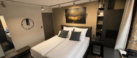 A hotel room with a bed, bedside table, a clock, a painting, a desk, and a chair.