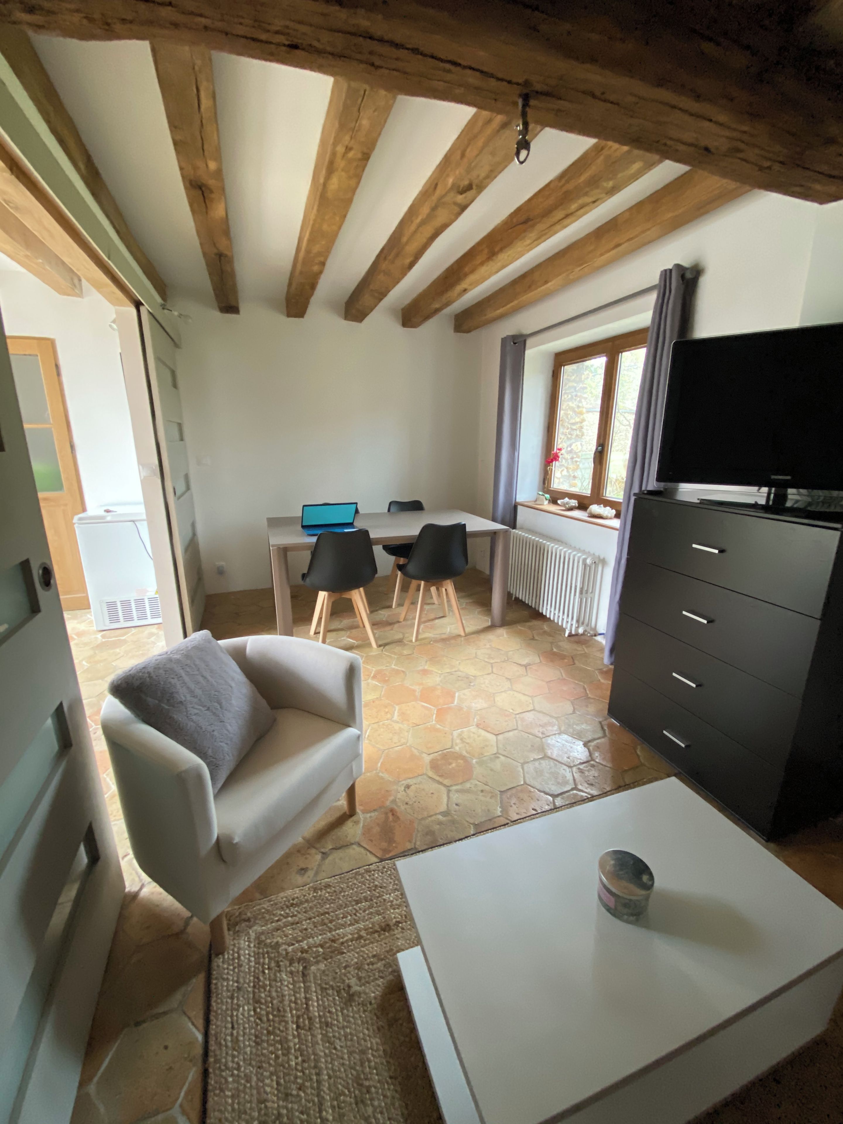 Una habitación con un sofá blanco, una mesita, un televisor sobre un mueble bajo, un escritorio con una computadora portátil y una ventana con cortinas.