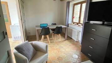 Una habitación con un sofá blanco, una mesita, un televisor sobre un mueble bajo, un escritorio con una computadora portátil y una ventana con cortinas.