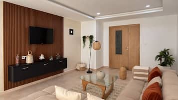 Een moderne woonkamer met een bank, een glazen salontafel, een televisie bevestigd aan een houten wandpaneel, een houten deur en een kamerplant.