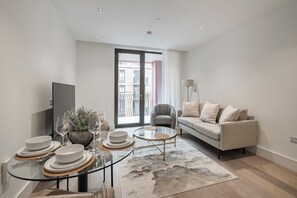 A modern living room with a sofa, a glass coffee table, and a dining area with plates and glasses. - Elegant One-Bedroom Retreat (London)