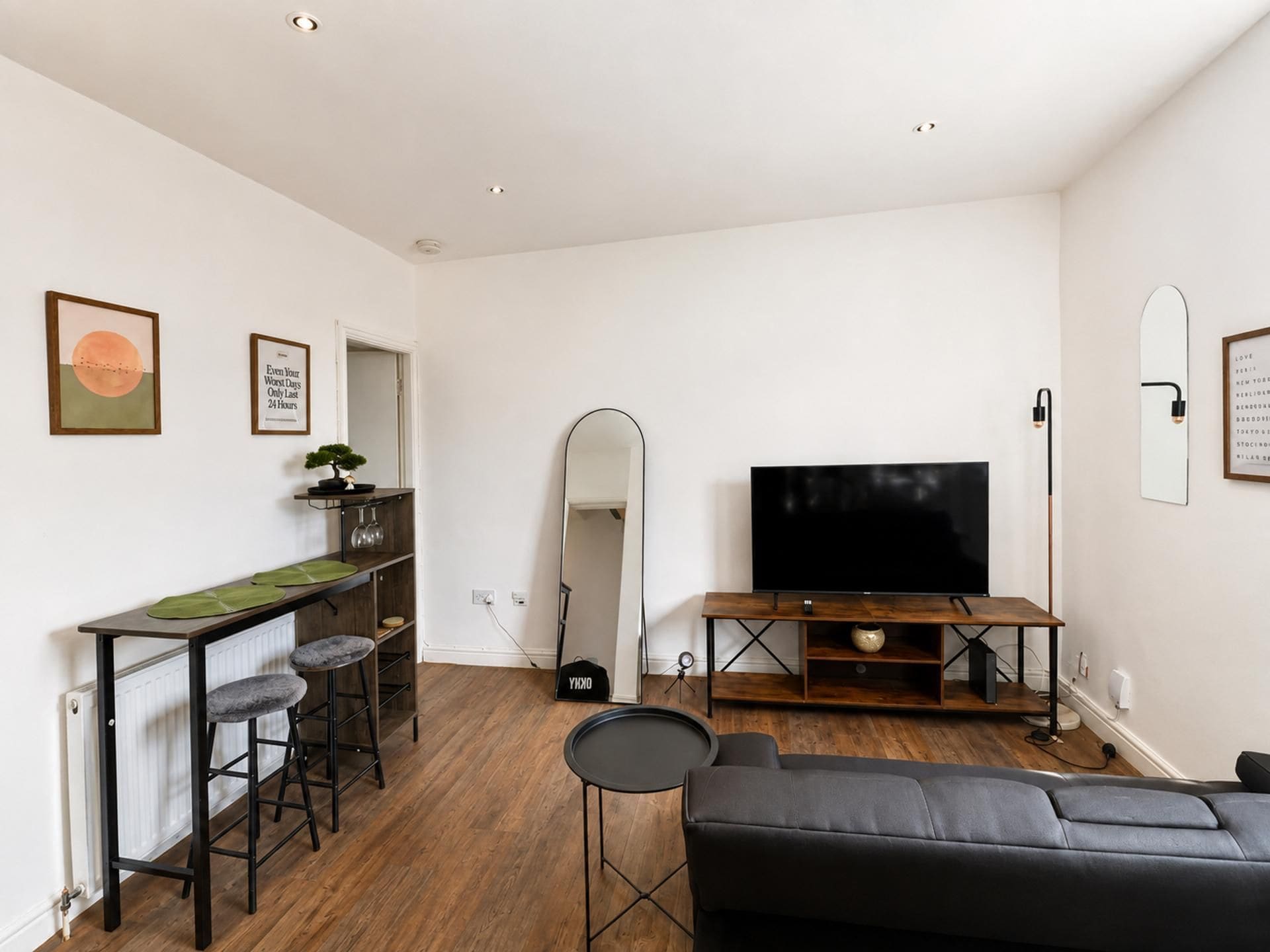 A modern living room with a black sofa, a TV on a wooden stand, a dining area with stools, and a large mirror.